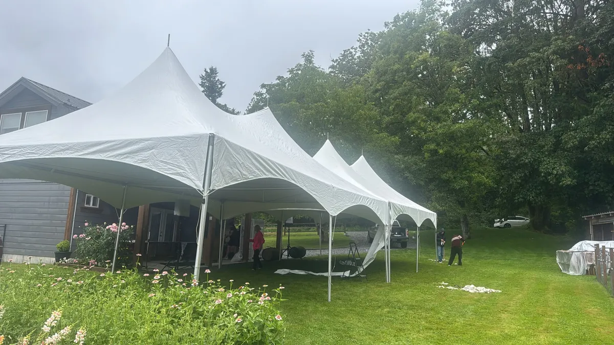 Three joined white high-peak tents beside a backyard garden, crew on site