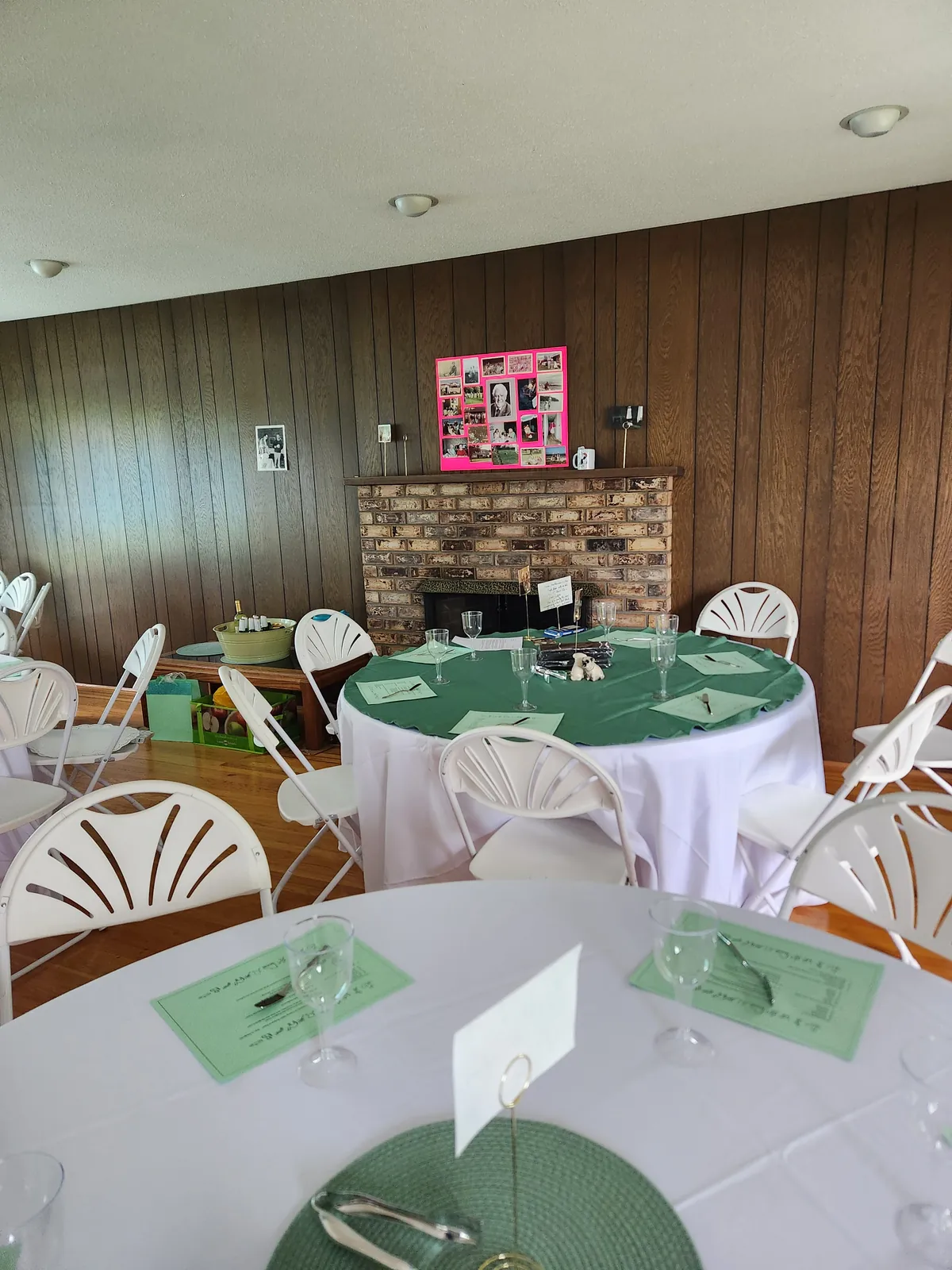 Round tables with white linens, green overlays, and white fanback chairs in a wood-paneled room with a brick fireplace