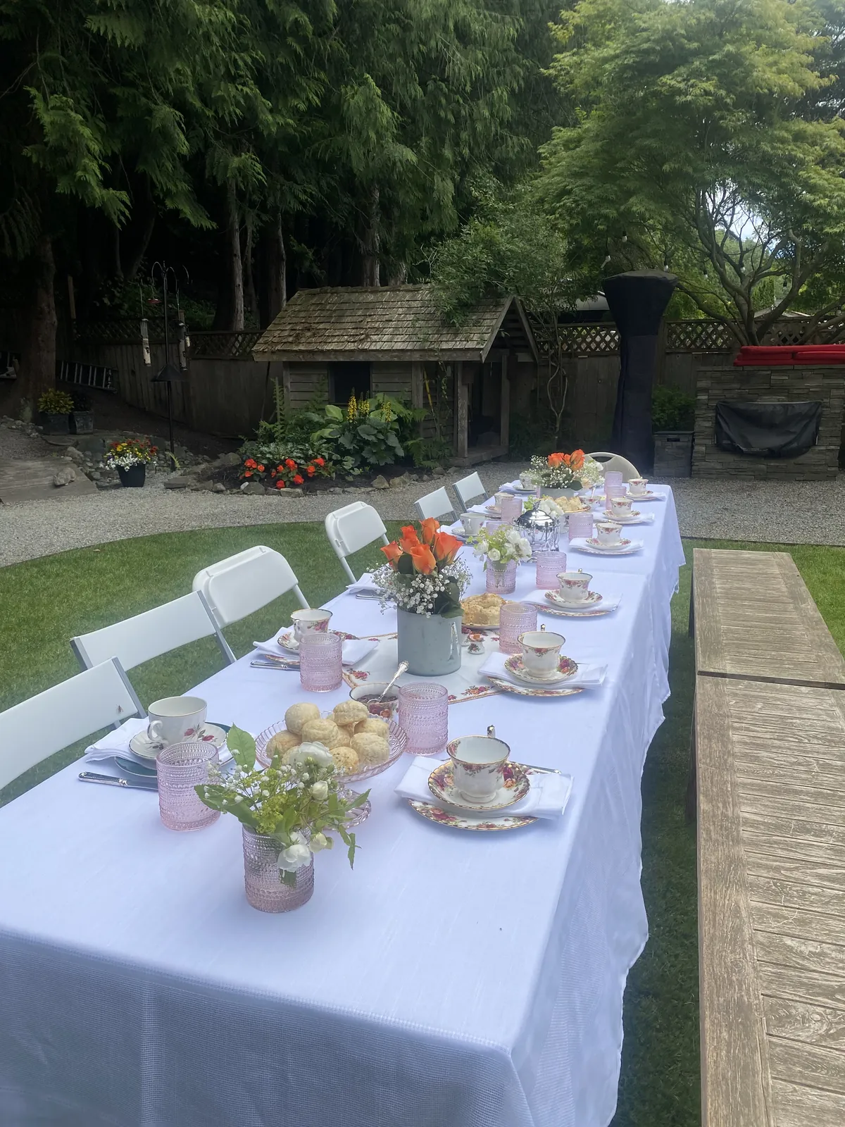 A long garden table with white folding chairs, floral teacups, and a cedar hedge behind on a North Vancouver job