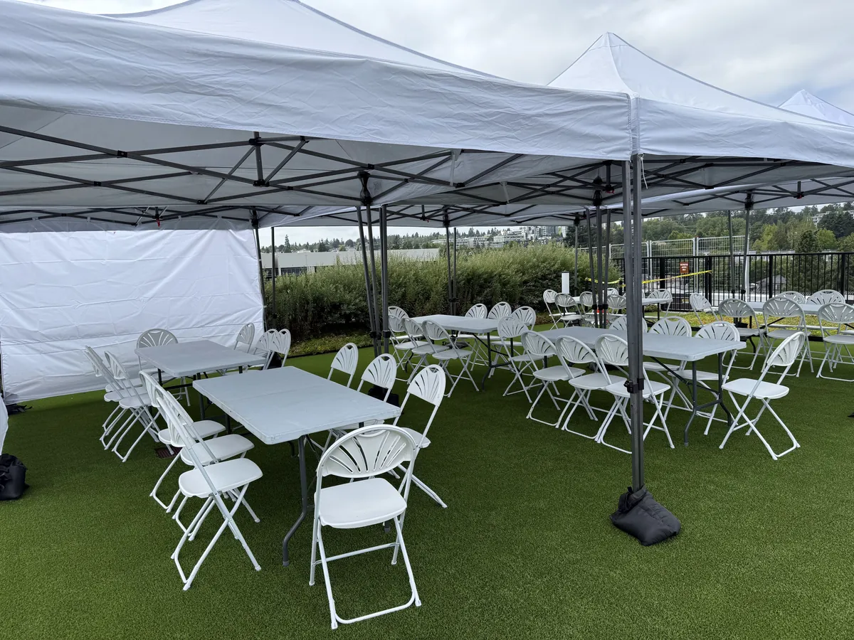 Joined pop-up canopies with banquet tables and fanback chairs on artificial turf
