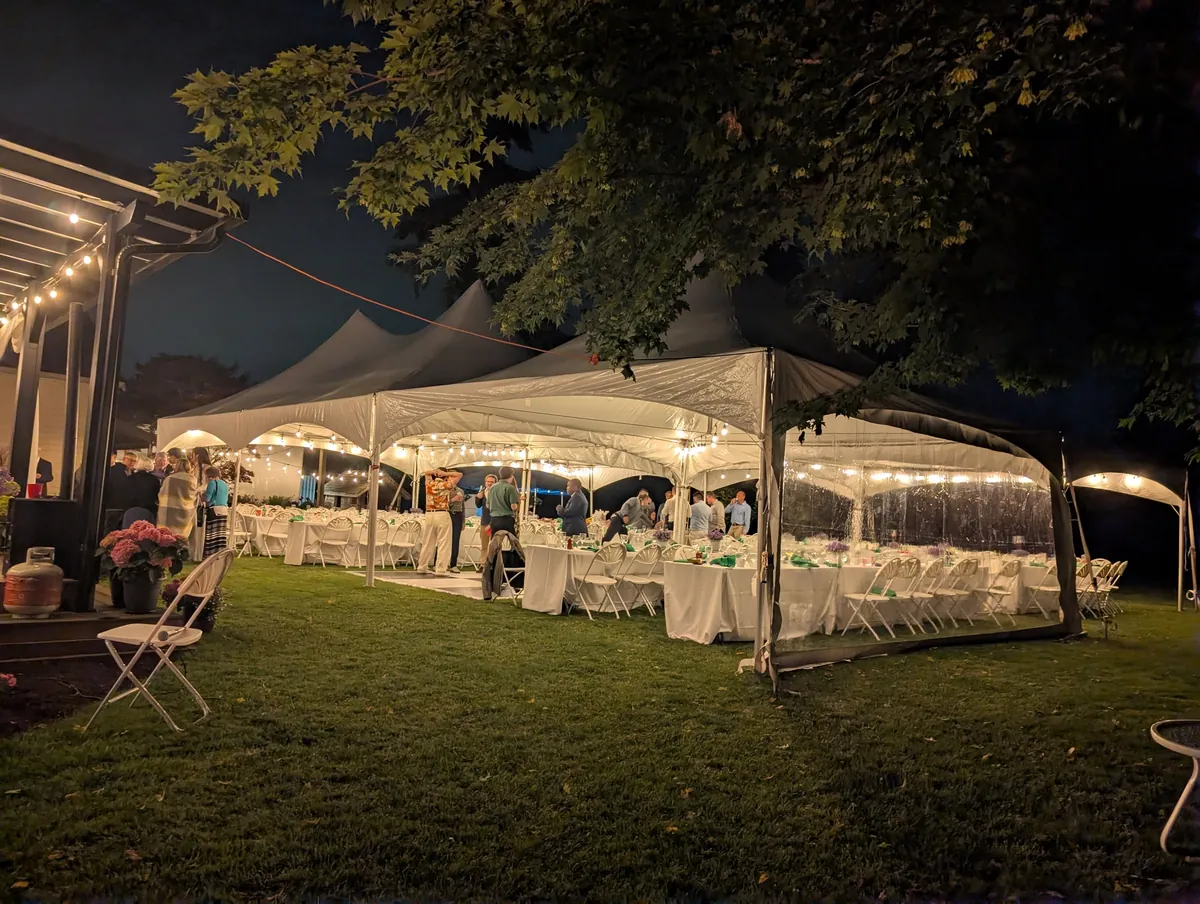 A backyard wedding reception at night under connected tents with string lights and clear sidewalls glowing