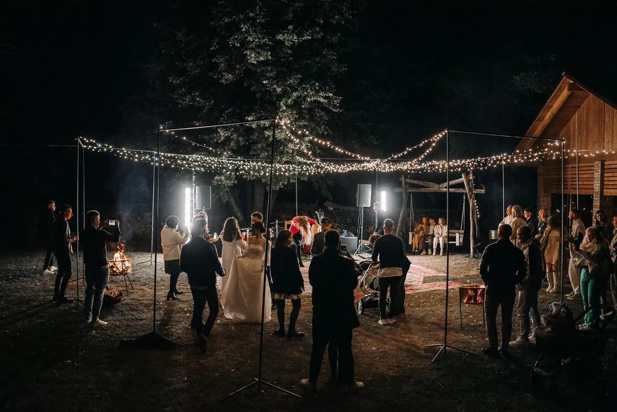 Newlyweds and guests at an outdoor reception lit by warm string lights at night