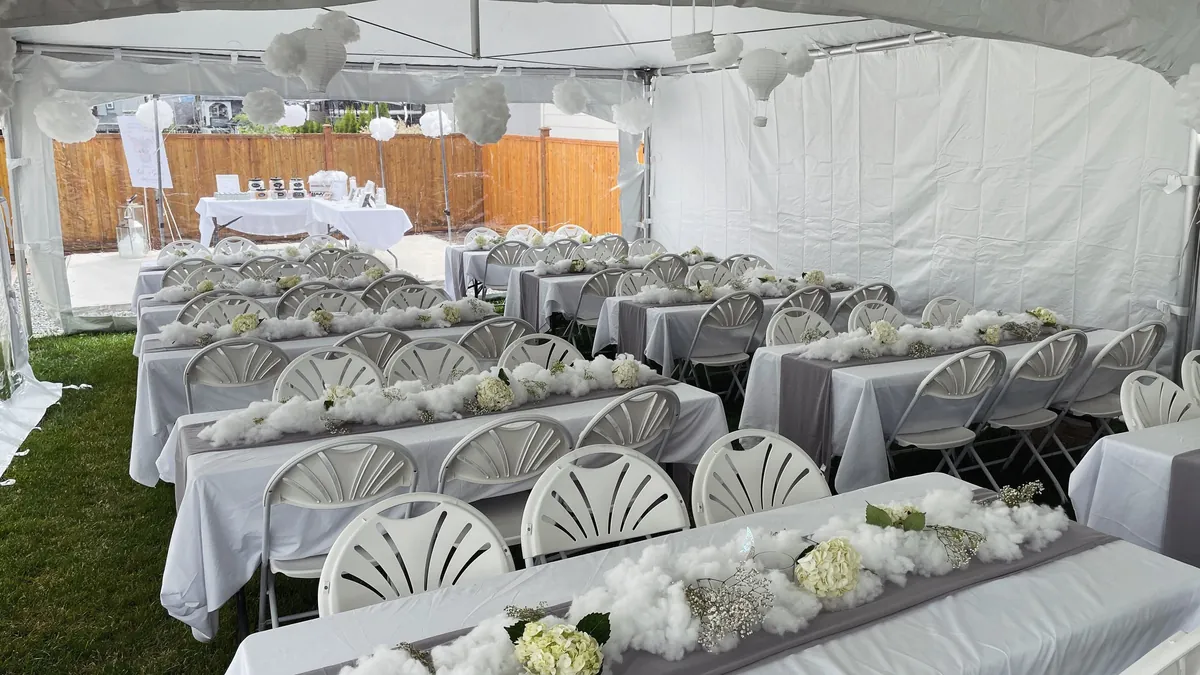Banquet tables with white linens and fanback folding chairs set for a reception inside a frame tent