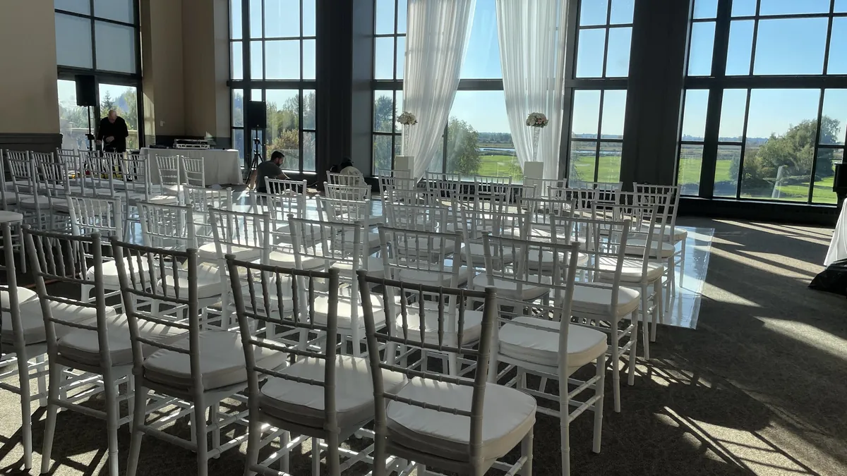 Rows of white Chiavari chairs set for a wedding ceremony in a bright venue with floor-to-ceiling windows